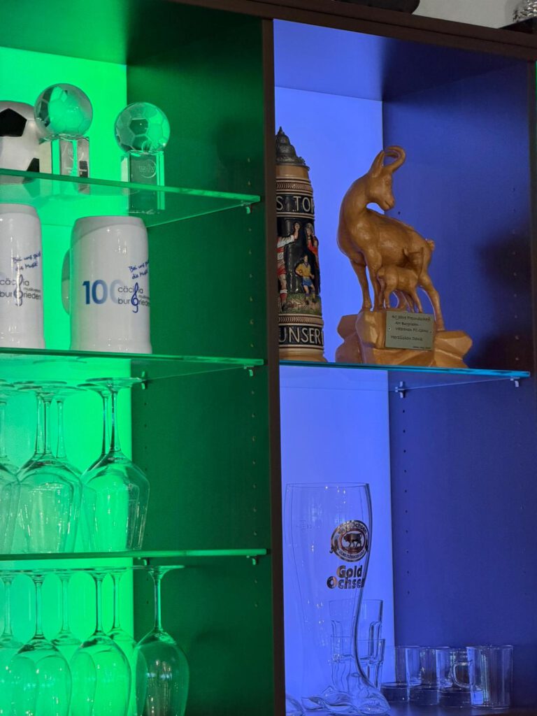 Regale mit verschiedenen Glaesern und dekorativen Objekten, beleuchtet in gruen und blau.