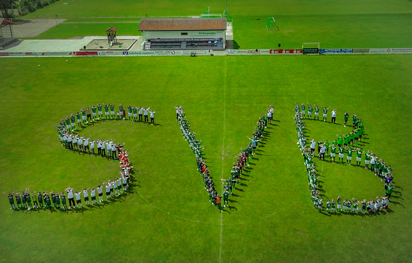 Luftaufnahme von Menschen, die auf einem Sportplatz die Buchstaben "SVB" formen.