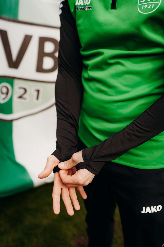 Eine Person in gruener Trainingsjacke vor einem Vereinsbanner.