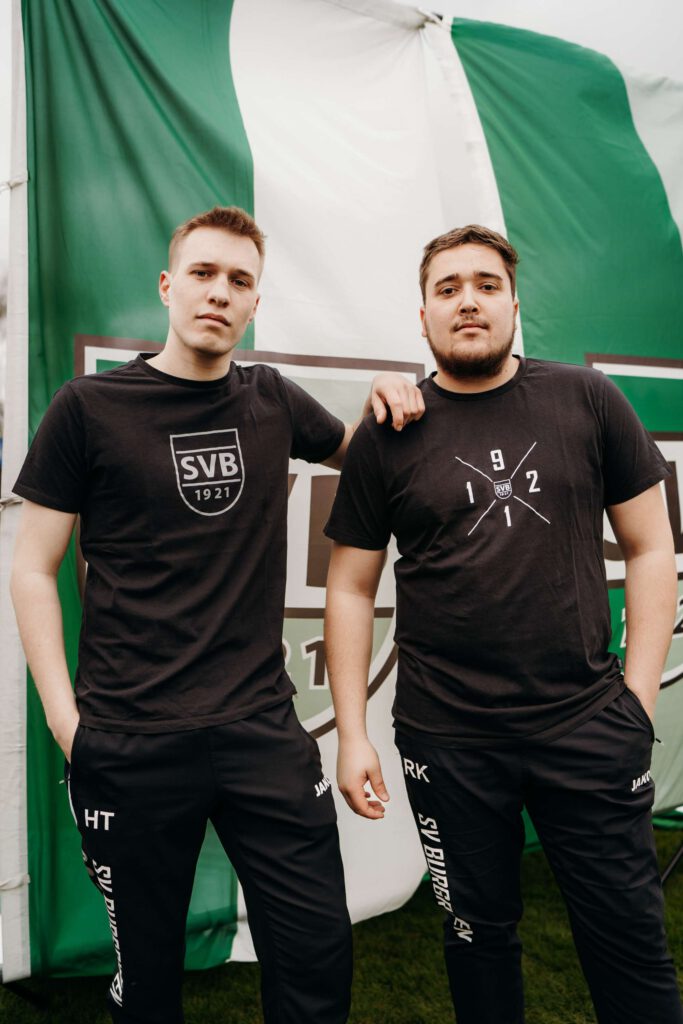 Zwei Maenner in schwarzen T-Shirts stehen vor einer gruenen und weißen Fahne.