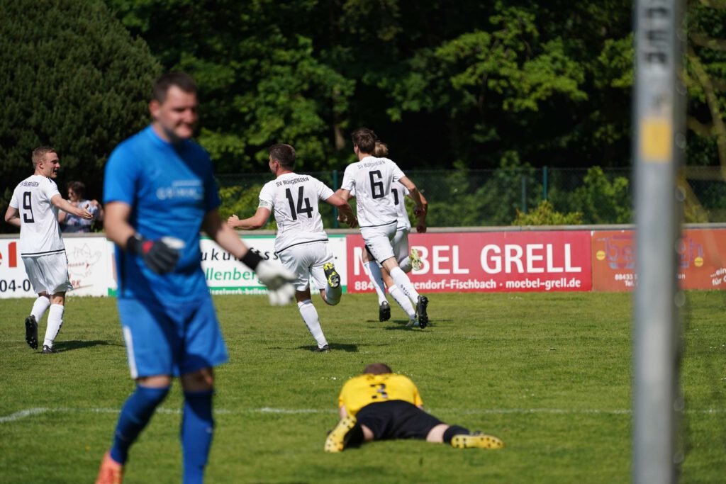 Ein Fußballspieler feiert ein Tor waehrend ein anderer auf dem Feld liegt.