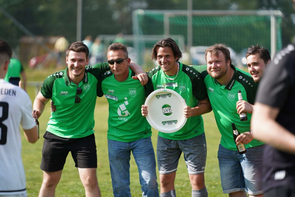 Eine froehliche Fußballmannschaft in gruenen Trikots posiert auf dem Spielfeld mit einem Tellersymbol in der Hand.