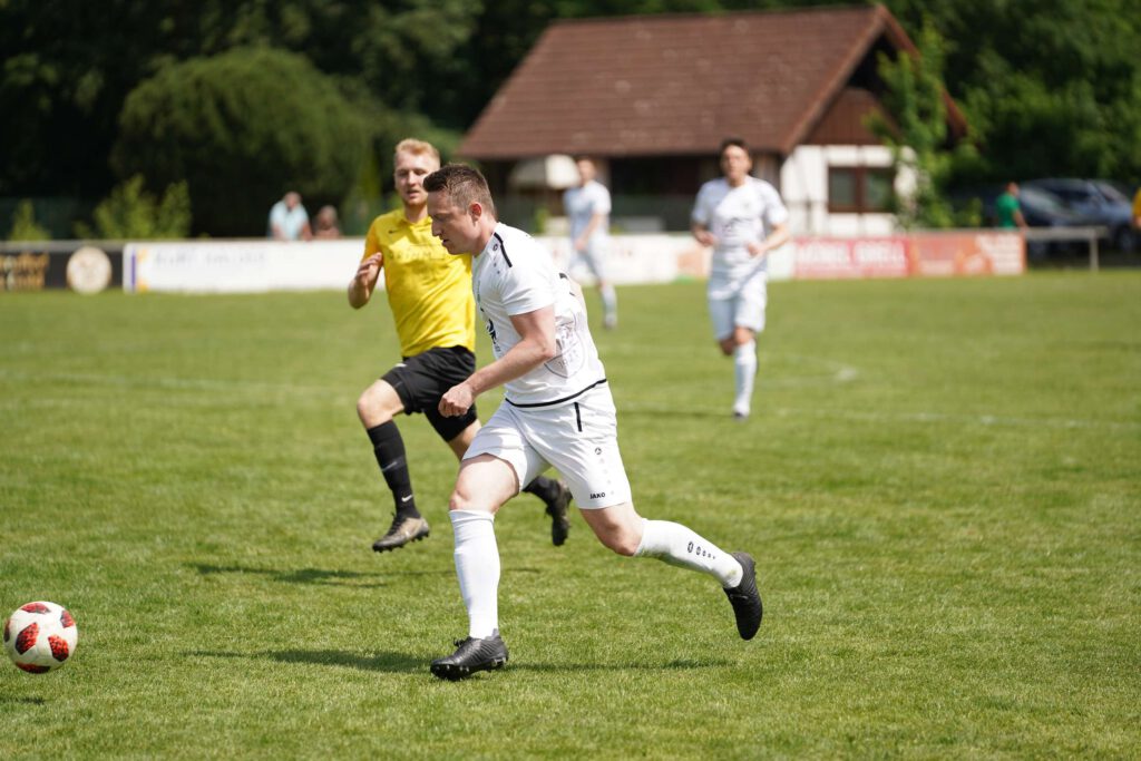Ein Fußballspieler in weißem Trikot rennt mit dem Ball vor einem Spieler in gelbem Trikot auf einem Rasenfeld.