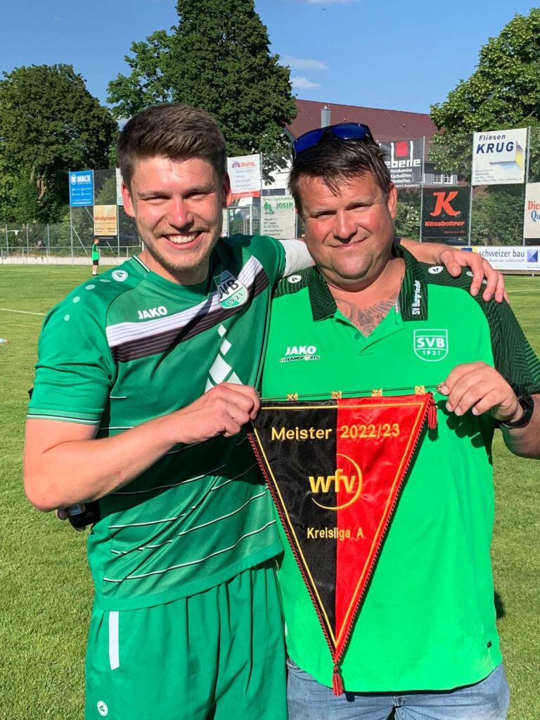 Zwei Personen in gruenen Trikots halten einen Wimpel auf einem Fußballplatz.