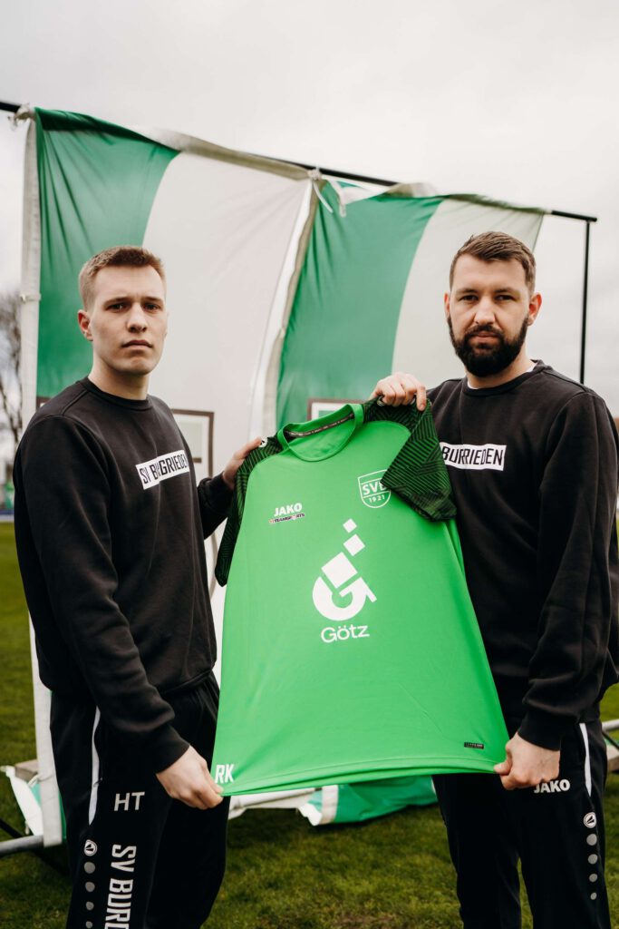 Zwei Maenner praesentieren ein gruenes Fußballtrikot vor einem Stadionhintergrund.