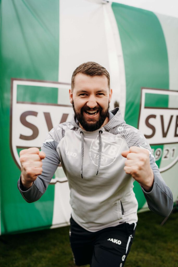 Ein Fußballtrainer jubelt enthusiastisch vor einer gruenen Werbetafel.