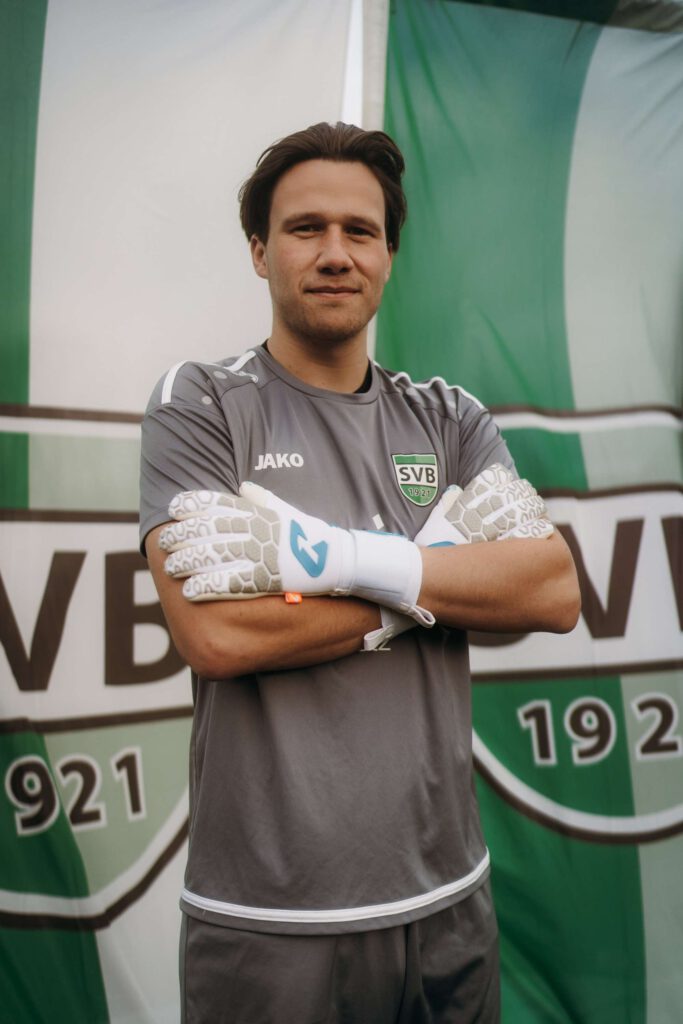 Ein Torhueter steht vor einem Fußball-Banner mit verschraenkten Armen.