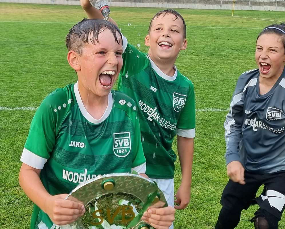 Kinder feiern auf dem Fußballfeld mit einem Pokal und gruenen Luftballons.