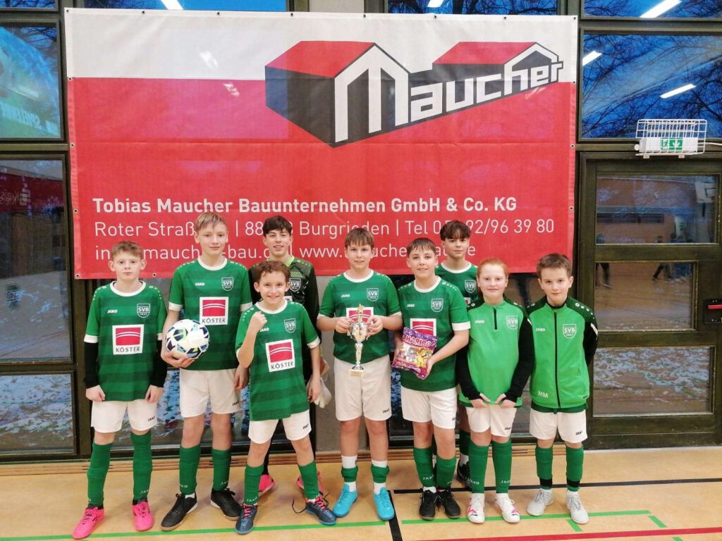 Eine Kinderfußballmannschaft posiert in gruenen Trikots vor einem Banner in einer Sporthalle.