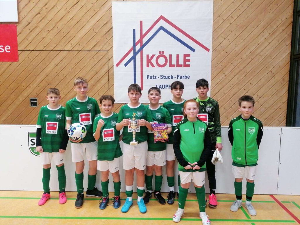 Eine Jugendfußballmannschaft in gruenen Trikots posiert in der Sporthalle vor einem Koelle-Banner.