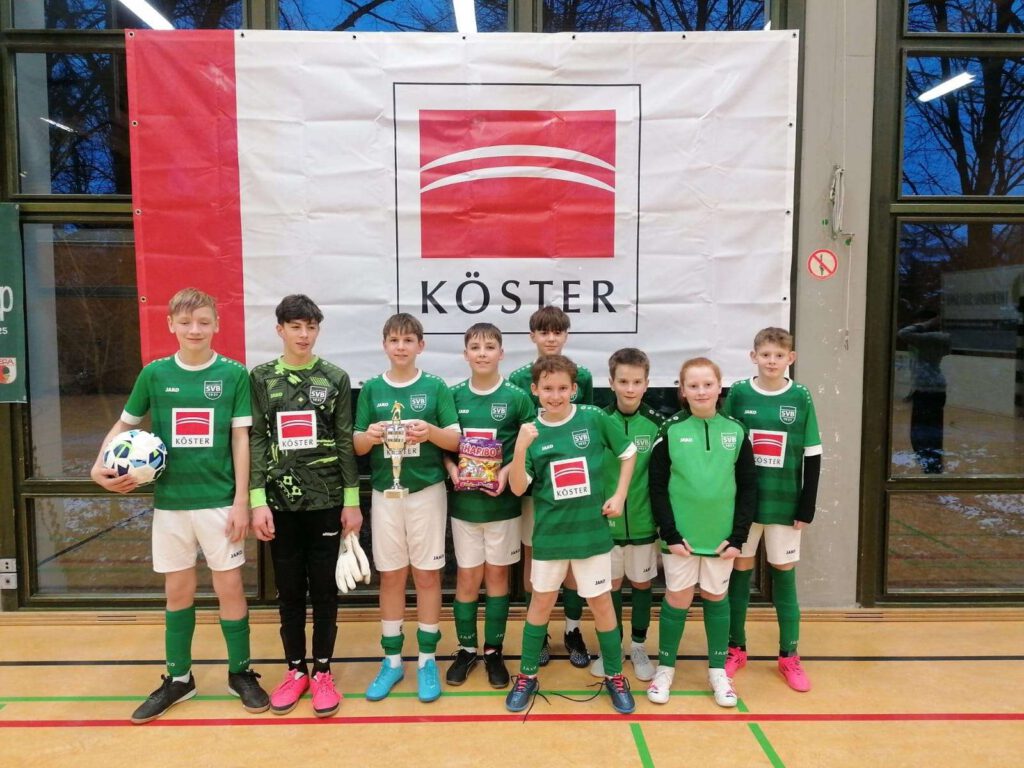 Eine Gruppe von jungen Fußballern in gruenen Trikots posiert fuer ein Mannschaftsfoto in einer Sporthalle.