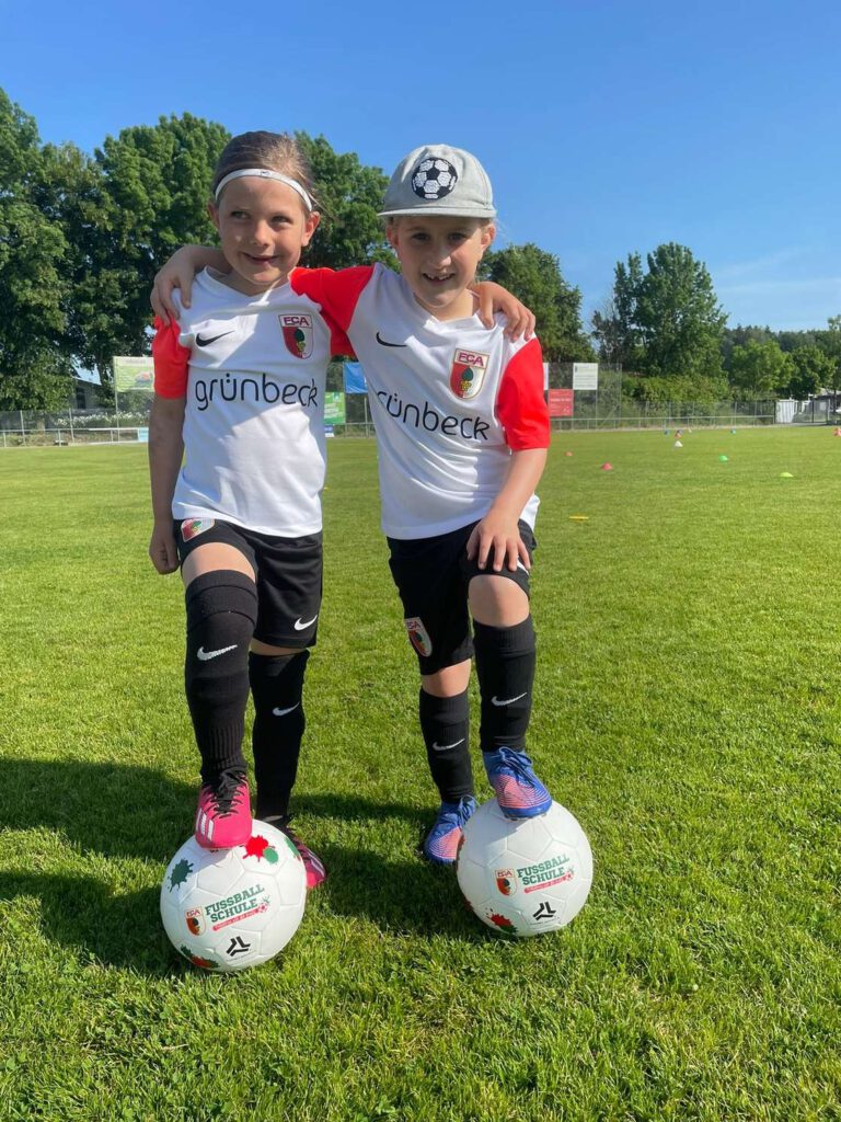 Zwei Kinder in Fußballtrikots stehen auf einem Spielfeld und posieren mit Fußballen auf dem Rasen.