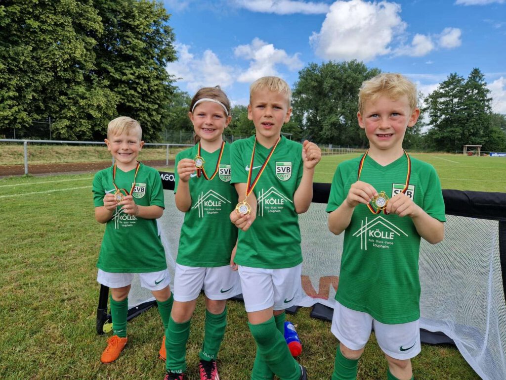 Eine Gruppe von vier Kindern in gruenen Fußballtrikots steht auf einem Fußballfeld und laechelt in die Kamera.