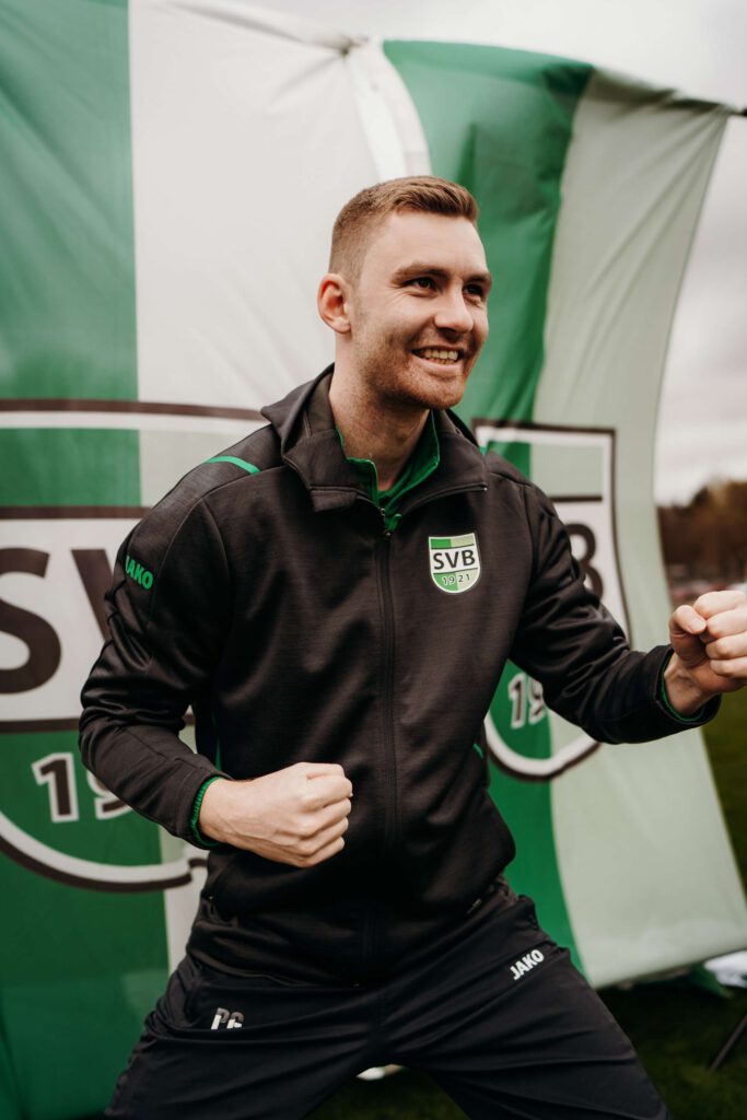 Ein sportlicher Mann in Trainingskleidung posiert enthusiastisch vor einem Vereinslogo.