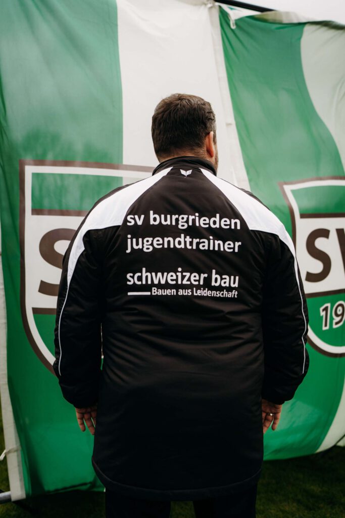 Person mit Werbeaufdruck auf Jacke vor einem gruenen Banner mit Vereinslogo