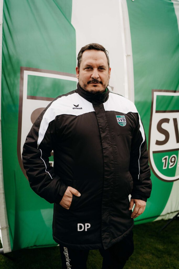 Person in einer Vereinsjacke steht vor einem großen Vereinslogo.